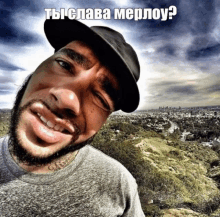 a man with a beard wearing a hat and a gray shirt is smiling in front of a landscape