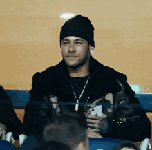 a man wearing a beanie and a camouflage jacket is sitting in the stands holding a cell phone .