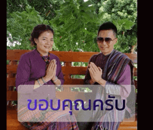 a man and a woman are sitting on a bench with their hands folded in front of a sign that says ขอบคุณ ครับ