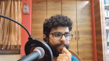 a man with glasses and a beard is sitting in front of a microphone with a clock on the wall behind him