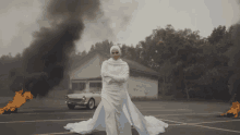 a woman in a white dress stands in a parking lot with a car on fire in the background