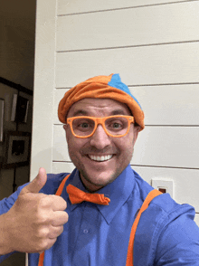 a man in a blue shirt and orange suspenders is wearing a hat and glasses and giving a thumbs up