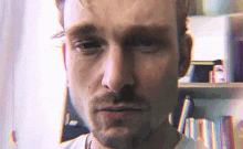 a close up of a man 's face with a beard looking at the camera in front of a bookshelf .