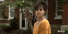 a woman in a yellow shirt is standing in front of a brick house with netflix written on the bottom right