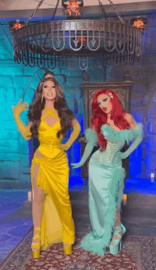 two drag queens dressed as princesses are standing next to each other in front of a chandelier .