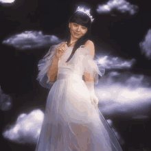 a woman in a white dress and tiara stands in front of a cloudy sky