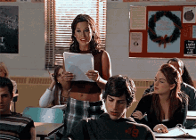 a woman stands in front of a group of students in a classroom with a bulletin board that says " happy holidays " on it