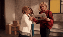 a woman holding a newspaper talks to another woman in front of a stack of boxes that say ' moving '