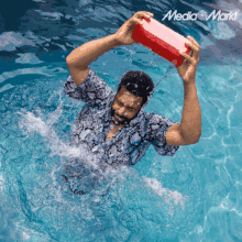 a man in a swimming pool is holding a red box over his head with the media markt logo in the corner