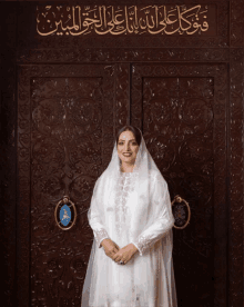 a woman in a white dress stands in front of a door with arabic writing