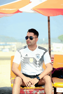 a man wearing sunglasses and an adidas shirt sits under an orange umbrella