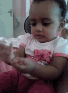 a baby girl in a pink and white shirt is sitting on a bed and playing with her hands .