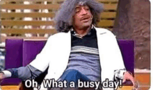 a man with a wig is sitting on a couch and saying `` oh what a busy day ! ''