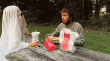 a man in a ghost costume is sitting at a picnic table with a bag of mcdonald 's french fries