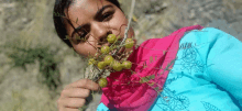 a woman wearing a pink scarf and a blue shirt holds a bunch of berries in her mouth