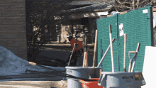 a man in an orange shirt is standing in front of a green fence with buckets and shovels in front of it