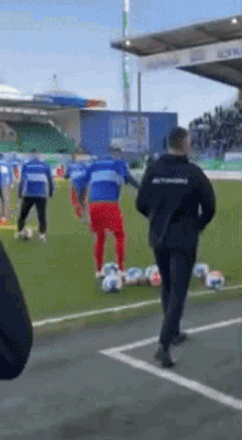 a man in a black jacket is walking on a soccer field with a bunch of soccer balls on the ground