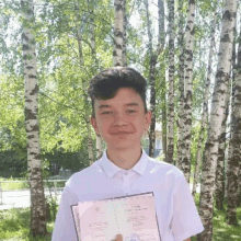 a young man in a white shirt is holding a diploma in front of a forest of birch trees .