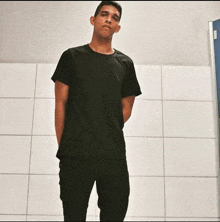 a young man wearing a black shirt and black pants stands in front of a white tiled wall