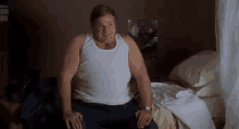 a fat man in a white tank top is sitting on a bed and smiling .