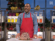 a man in a red apron stands in front of a cake with a face on it and a sign that says snl