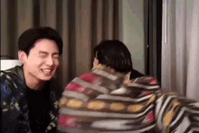 two men are sitting next to each other in a room and laughing . one of the men is wrapped in a blanket .