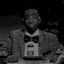 a young boy wearing glasses and a bow tie is sitting at a table with a plate of food .