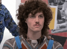 a man with curly hair and a moustache is wearing overalls and a sweater