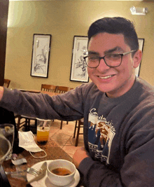 a young man wearing glasses and a sweater that says cordaches on it sits at a table with a bowl of soup