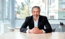 an older man is sitting at a table with his hands folded and smiling .