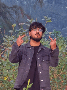 a man wearing a black shirt and a denim jacket stands in front of a bush giving the middle finger