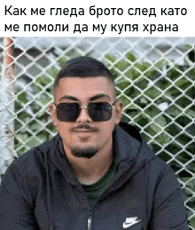 a man wearing sunglasses and a black jacket is sitting in front of a chain link fence .