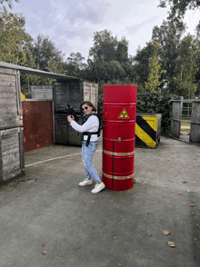 a woman holding a gun standing next to a red barrel with a yellow and black sign on it