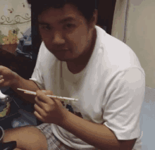 a man in a white shirt is sitting on a bed eating with chopsticks