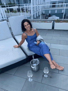 a woman in a blue jumpsuit sits on a lounge chair with a bucket of champagne and two glasses of wine