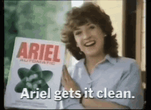 a woman is holding a box of ariel automatic laundry detergent and says ariel gets it clean .