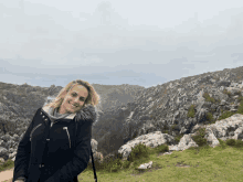 a woman in a black coat with a fur hood stands in front of a rocky landscape