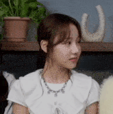 a young woman wearing a white shirt and a necklace is sitting in front of a shelf with potted plants .