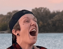 a man wearing a hat is laughing with his mouth open in front of a lake .