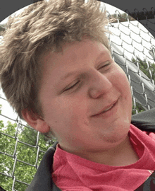 a young boy wearing a pink shirt is smiling with his eyes closed in front of a chain link fence