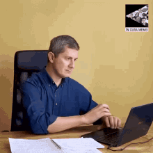 a man sits at a desk using a laptop with a picture of coins behind him that says in gura memei