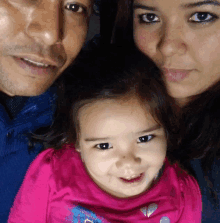 a little girl wearing a pink shirt with a heart on it smiles with her parents