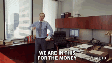 a man in a suit and tie is standing in front of a desk that says " we are in this for the money "