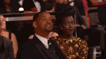 a man in a suit and tie sits next to a woman in a sequined dress while watching a live abc broadcast