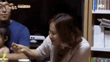 a woman is feeding a child with a spoon while sitting at a table in front of a bookshelf .