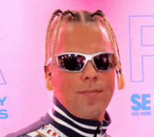 a man with braids wearing sunglasses stands in front of a pink background