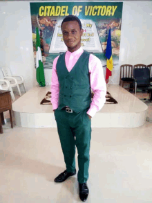 a man in a pink shirt and green vest stands in front of a sign that says citadel of victory