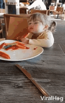 a little girl sits at a table with a plate of carrots and a knife with viralhog written on the bottom