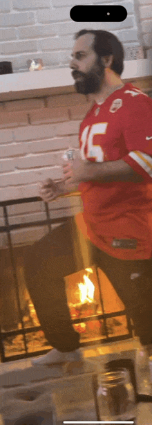 a man wearing a kansas city chiefs jersey is standing in front of a fireplace holding a beer