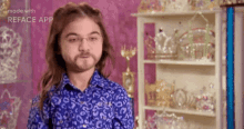 a man with a beard wearing glasses and a blue shirt is standing in front of a shelf full of tiaras .